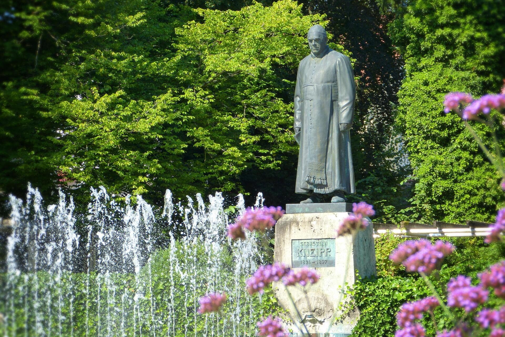 Statue von Sebastian Kneipp vor Springbrunnen und grünen Bäumen