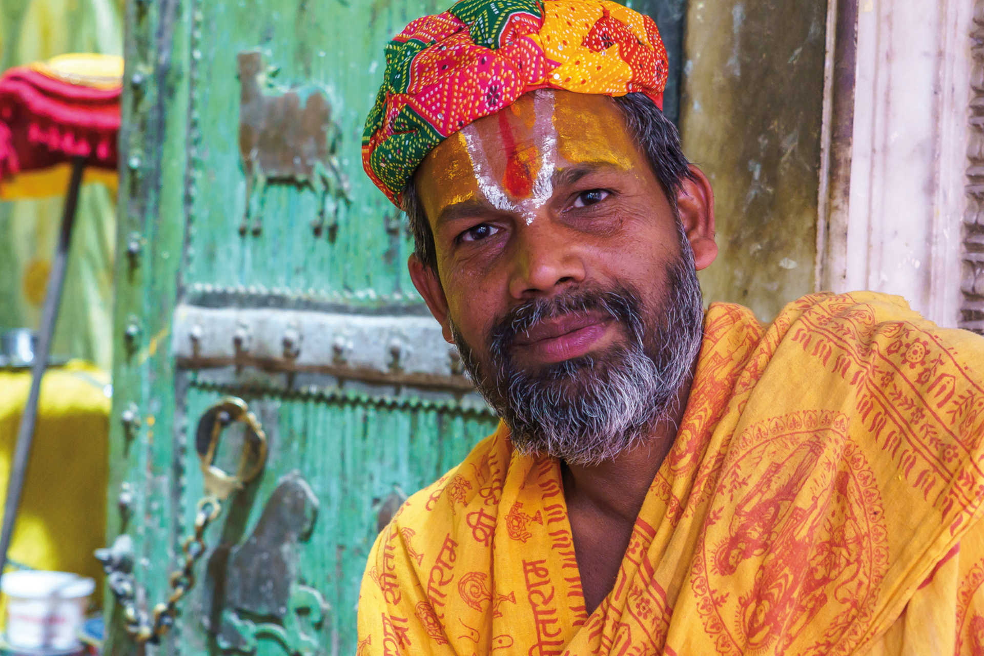 Mann mit Tilaka-Zeichen, gelbem Gewand mit Sanskrit und bunter Turban vor Tempel