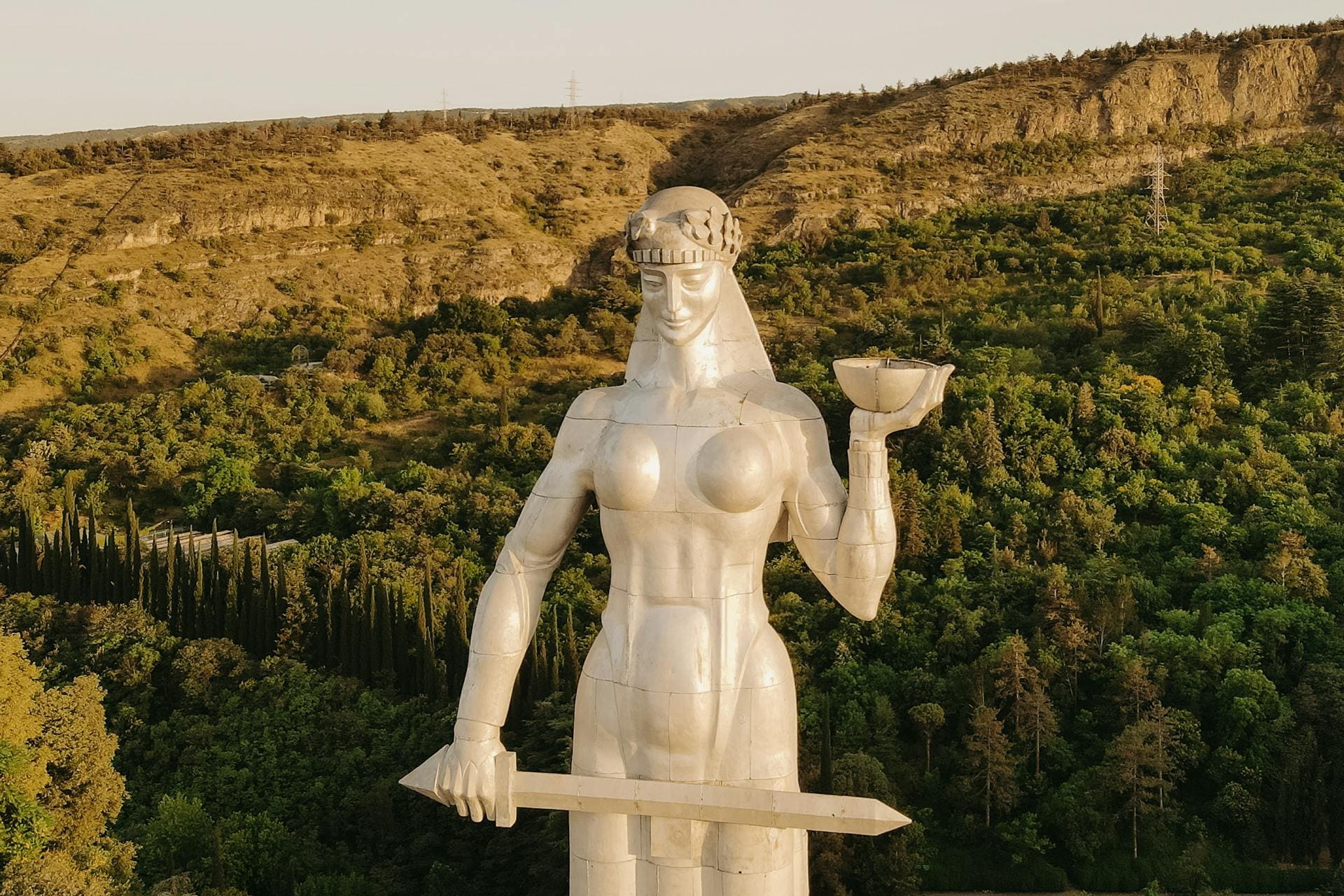 Große silberne Statue mit Schwert und Schale vor bewaldeten Hügeln