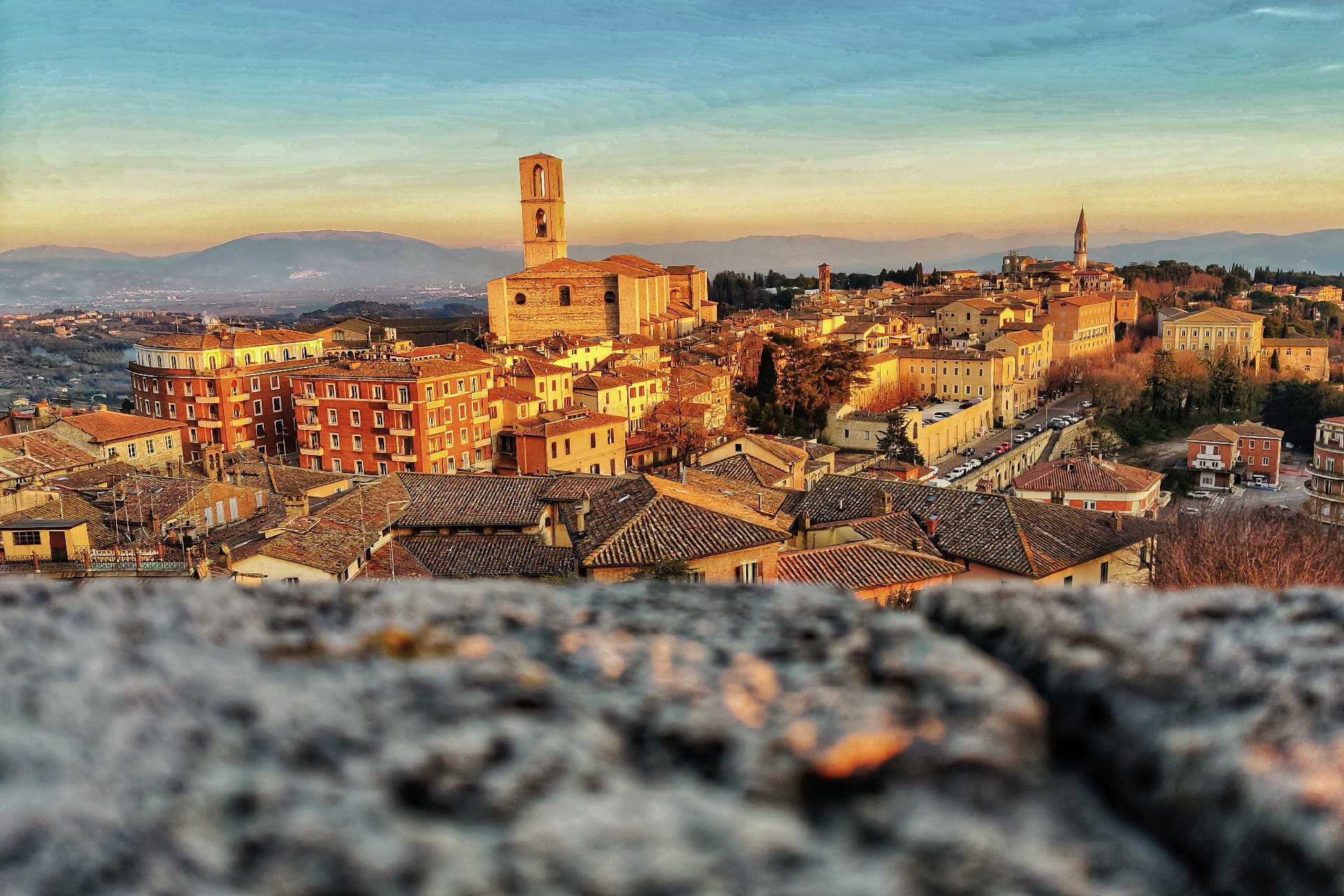 Hügelige Stadtlandschaft mit Kirchen und warm beleuchteten Häusern bei Sonnenuntergang