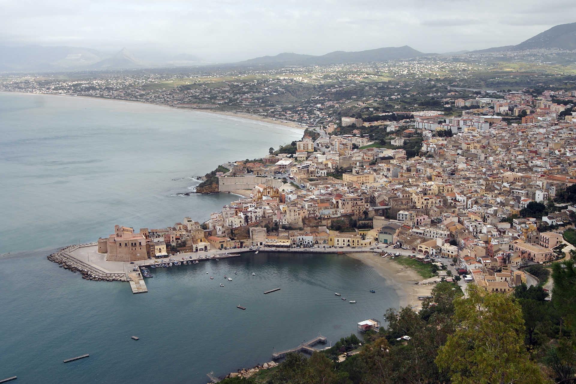 Blick auf die sizilianische Küste mit der historischen Altstadt von Castellammare del Golfo und der markanten Festung am Hafen, eingerahmt von sanften Hügeln und dem weiten Mittelmeer.