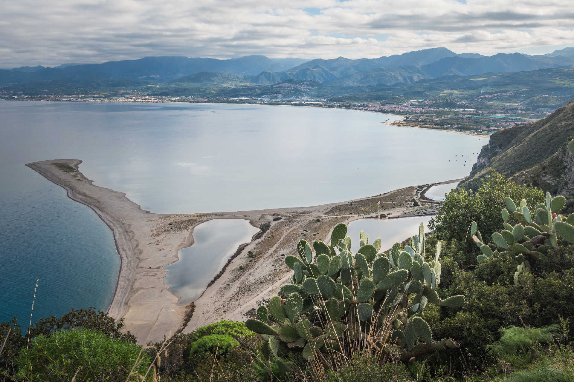 Blick auf die sichelförmige Sandbank von Tindari an der Nordküste Siziliens mit ruhiger See, Lagunen und grüner Küstenlandschaft im Hintergrund.