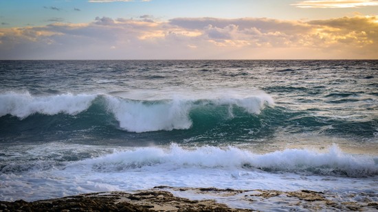Wellen brechen am Ufer unter bewölktem Himmel bei Sonnenuntergang